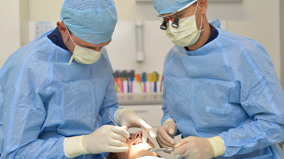 Dwóch dentystów w niebieskich fartuchach zapewnia opiekę periodontologiczną pacjentowi w Katowicach.