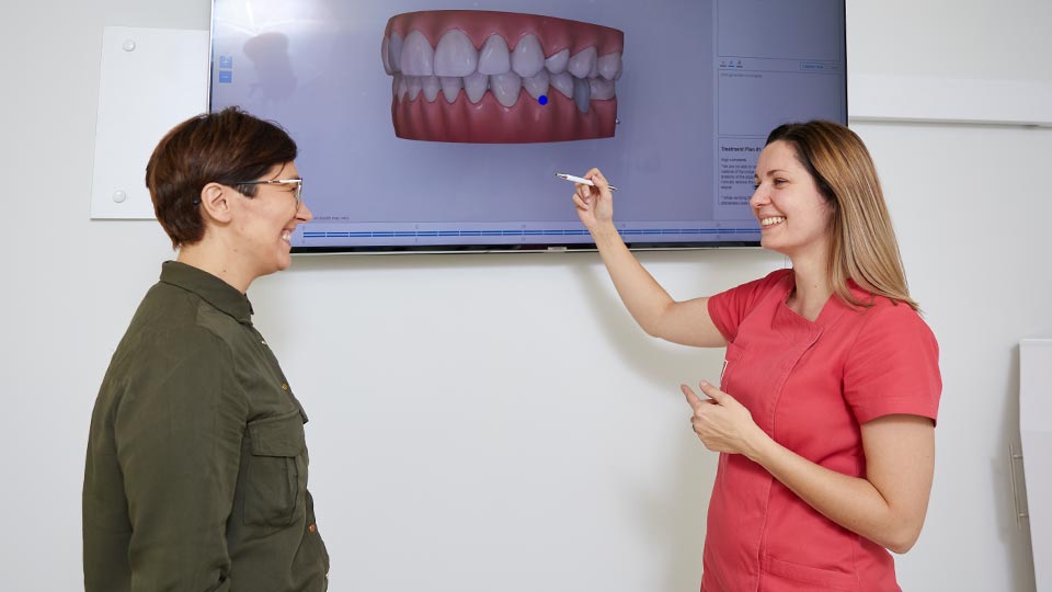 Dwie kobiety w gabinecie dentystycznym wskazujące na aparat na zęby na ekranie telewizora.
